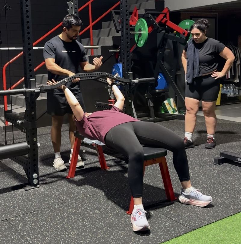 WSF coach guiding a member through a bench press while another member waits their turn